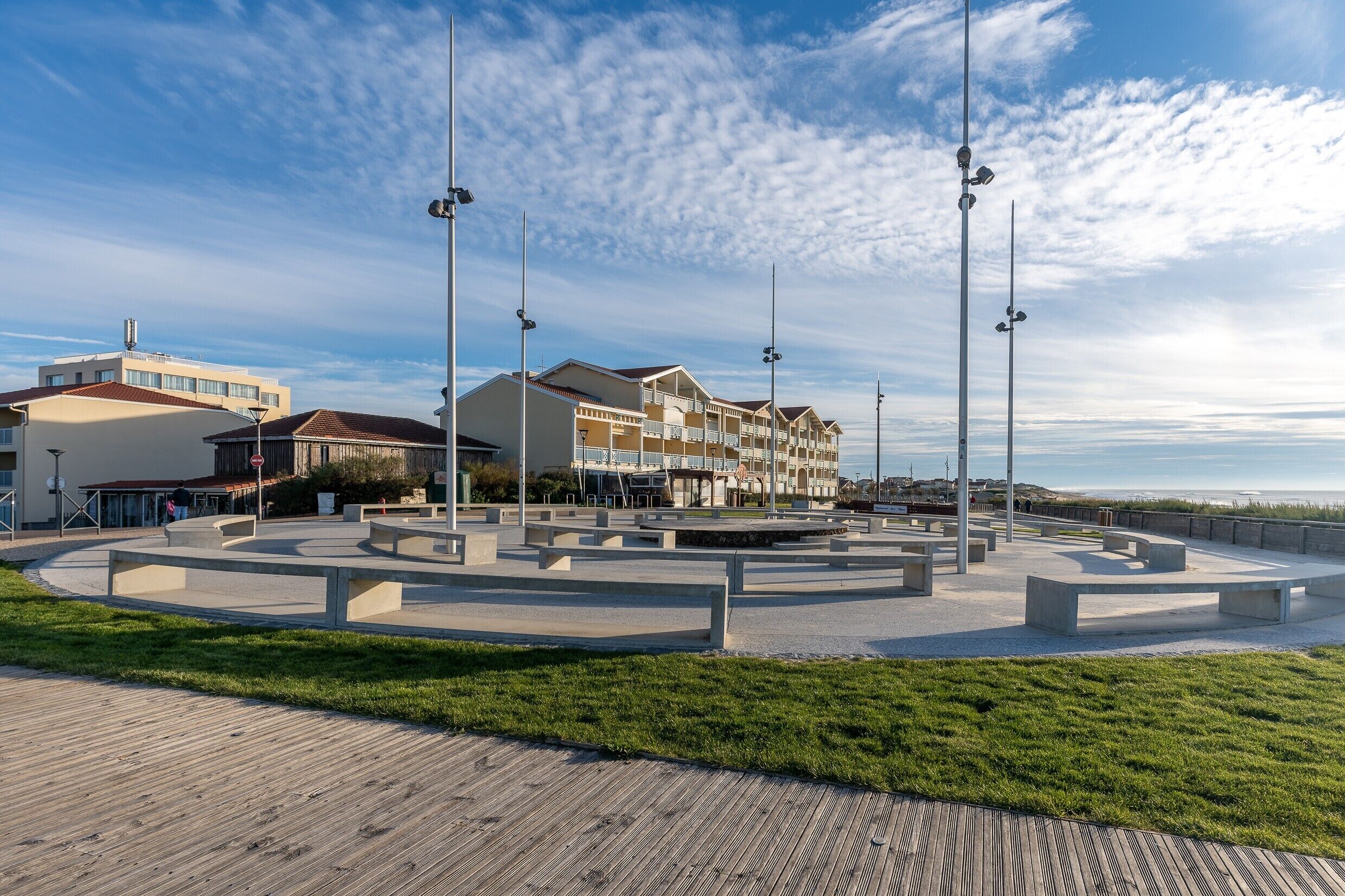 A coastal promenade with a circular seating area, street lamps, and a row of buildings with balconies.
