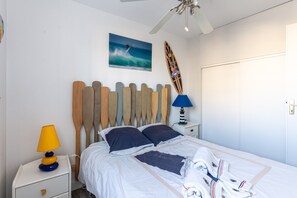 A bedroom with a bed, a wooden headboard, a nightstand with a lamp, a surfboard decoration, and a painting of a surfer on a wave. - Apartment "Didier" with Sea View, Terrace & Wi-Fi (Mimizan)