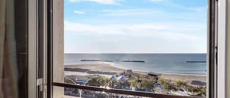 A view from a window overlooking a beach with a clear sky, a parking area, and a building with a blue roof.