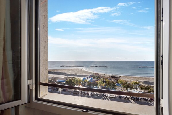 A view from a window overlooking a beach with a clear sky, a parking area, and a building with a blue roof.