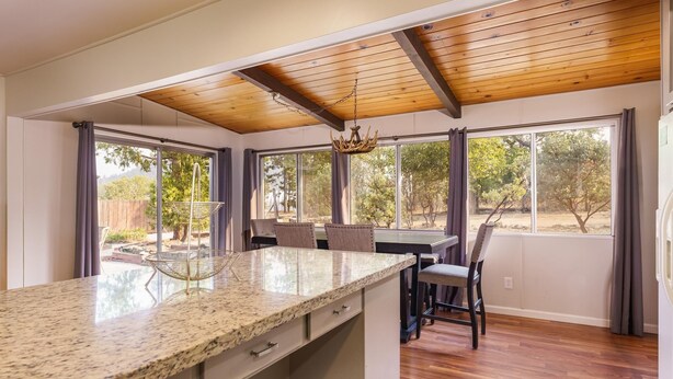 A kitchen with a granite countertop, wooden ceiling beams, and large windows with curtains. - Lake View Hideaway Near Yosemite & Bass Lake (Oakhurst)