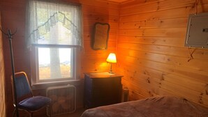 Acogedora habitación tipo cabaña con una cama, una silla, un escritorio con lámpara, un calefactor y una ventana con persianas.