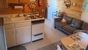 Cocina compacta con estufa, lavamanos y armarios de madera. Se ve un pequeño comedor con una mesa y un sofá.