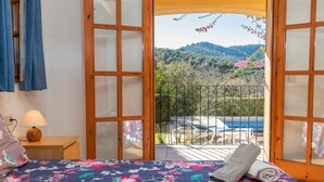 A bedroom with a bed, a nightstand, a lamp, and a view of a balcony with a railing and some plants. - Villa "Rosa" with Mountain View, Pool & Wi-Fi (Calonge)