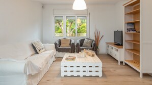 A living room with a white sofa, two armchairs, a coffee table, a TV on a wooden shelf, and a window with a view of trees. - Villa "Terrassa" close to the Beach with Pool & Wi-Fi (Calonge)
