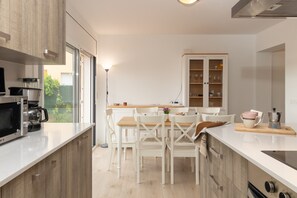 Una cocina moderna con armarios blancos, un comedor con sillas blancas, una cafetera y vistas a través de la puerta de vidrio.