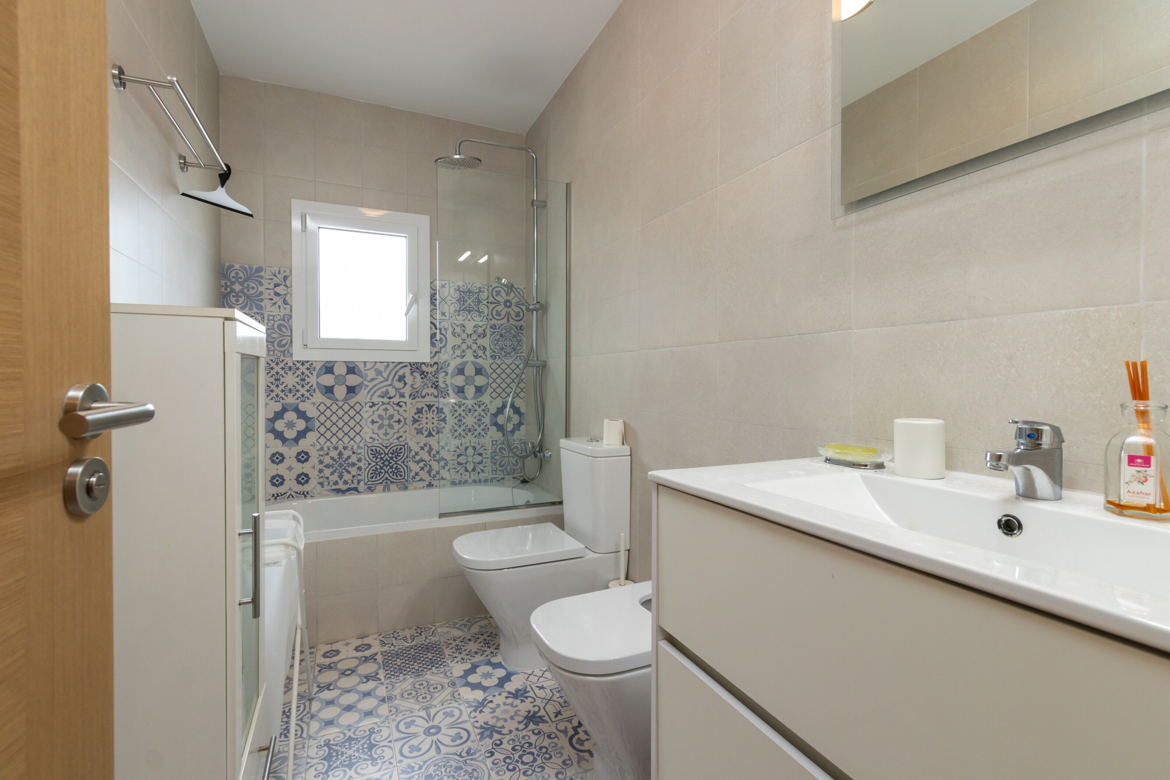 A bathroom with a shower, toilet, and sink, featuring patterned tiles and a window.