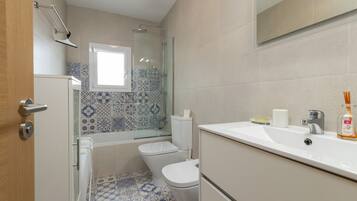 A bathroom with a shower, toilet, and sink, featuring patterned tiles and a window.