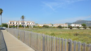 A coastal residential area with a wooden boardwalk, white buildings with red-tiled roofs, and a mountain in the background.