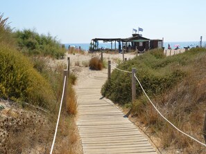 A wooden boardwalk leading to a beachside structure with flags, surrounded by vegetation.