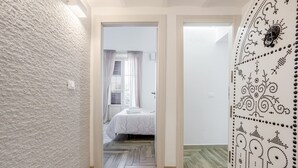 A hallway leading to a bedroom with a herringbone floor, a bed with a white duvet, and a door with a decorative pattern.