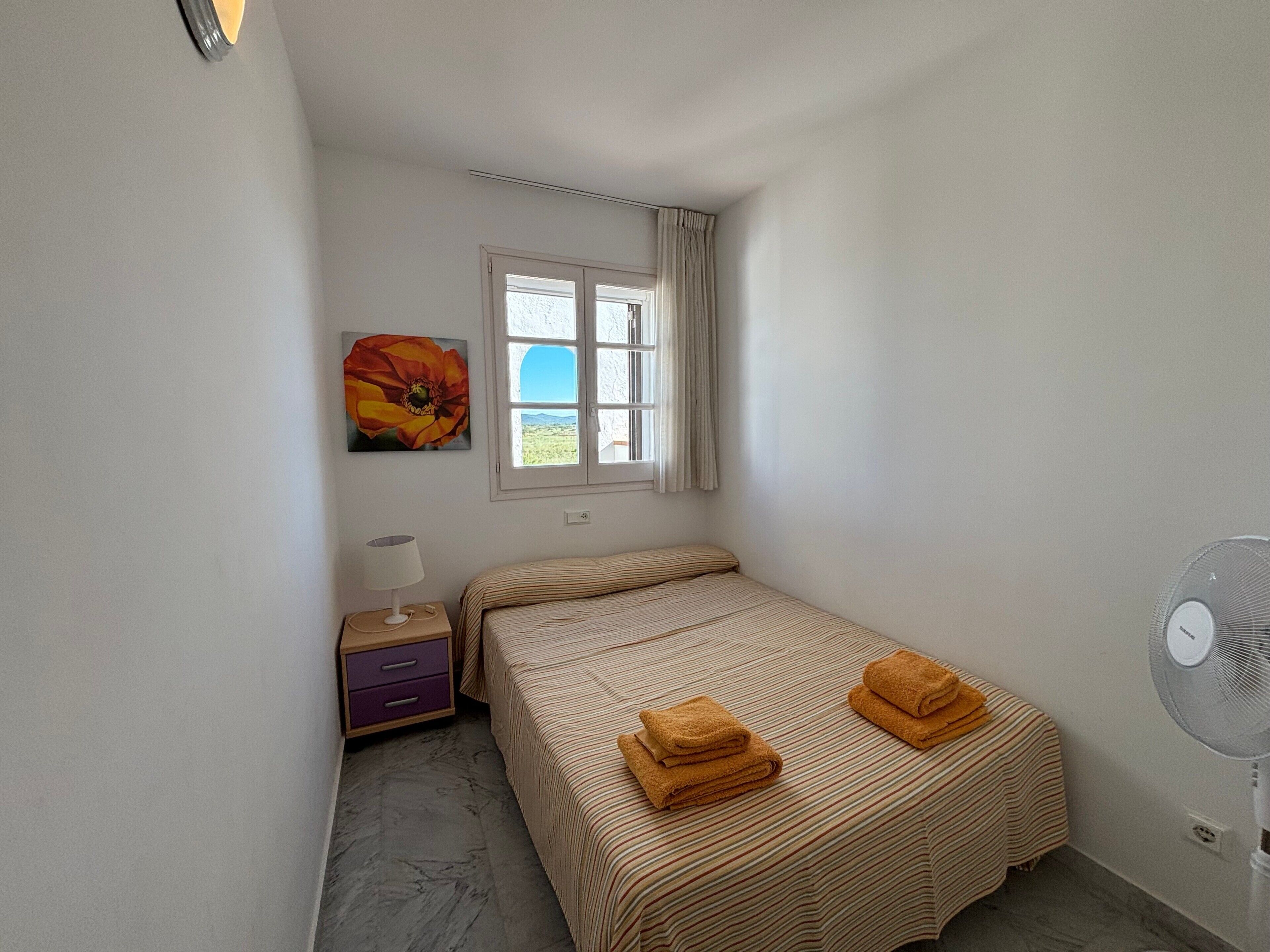 Quarto com cama, mesa de cabeceira, luminária, janela com cortinas, um quadro e um ventilador.