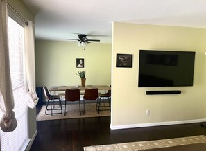 A living room with a dining area, a ceiling fan, a flat-screen TV mounted on the wall, and a potted plant.