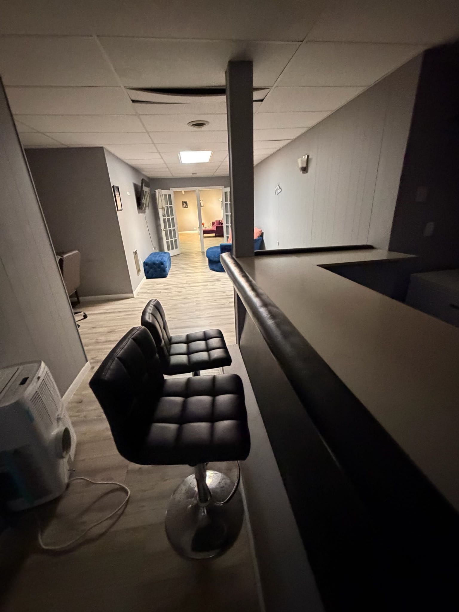 A hallway with a reception desk, two black chairs, a blue ottoman, and a white trash bin.