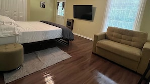 Ein Schlafzimmer mit Bett, Deckenventilator, Fernseher, Fenster mit Jalousien und einer Couch.