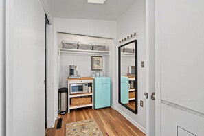 Enter a serene space with a compact hallway leading to a cozy bedroom, featuring a wardrobe, kitchenette with microwave and coffee maker, and a compact refrigerator.