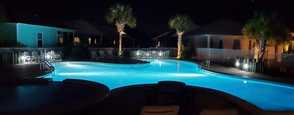 A nighttime swimming pool area with lounge chairs and palm trees, surrounded by buildings with illuminated windows.