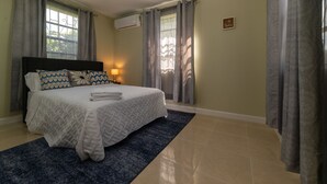 A neatly made bed with a white comforter and pillows, a blue area rug, and a wall-mounted air conditioning unit. - 3 Bedrooms in paradise w/fruit trees 3 mins-Oistins! (Oistins)