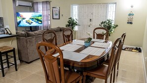 A dining area with a wooden table, chairs, and a TV in the background. - 3 Bedrooms in paradise w/fruit trees 3 mins-Oistins! (Oistins)