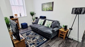 A living room with a grey sofa, a TV on a wooden stand, a patterned rug, and a painting of a forest.