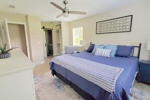 A bedroom with a bed, a ceiling fan, a bedside table, a lamp, a potted plant, and a door leading to another room.