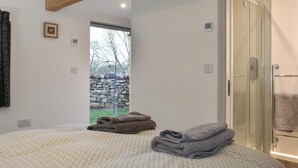 A bedroom with a bed, a wooden ceiling beam, a door leading to a bathroom, and a view of a stone wall and trees through a window.
