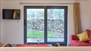 A room with a sliding glass door leading to a stone wall and garden, a flat-screen TV mounted on the wall, and a red couch with a yellow pillow.