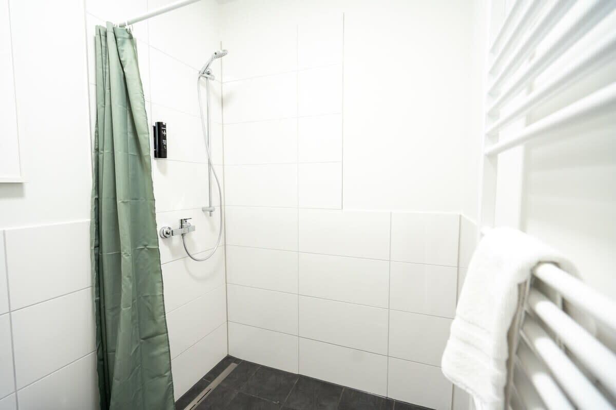 Een kleine, schone douche ruimte met een groen douche gordijn, witte tegels, en een witte handdoek op een radiator.