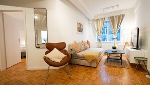 Una sala moderna con un sofá, una butaca, una mesita y una cama. Hay una ventana con cortinas, un espejo y una lámpara.