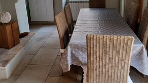 A dining area with a long table, wicker chairs, a hanging light fixture, and a view through a window.