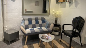 A modern living room with a sofa, a chair, a small table with magazines, a mirror, and a shelf with books and plants. - Studio 25 m² Côté Parc des Contades (Strasbourg)