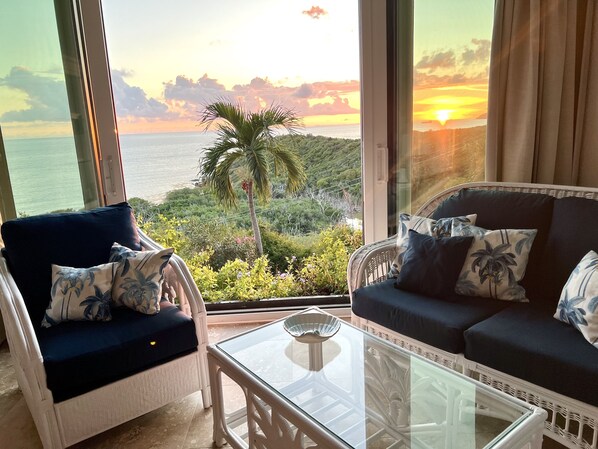 A balcony with a view of the sea and hills, furnished with a sofa and chairs, and a glass table.