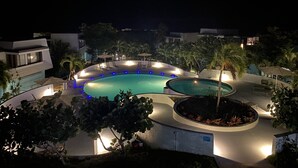 A nighttime view of a circular swimming pool with surrounding lounge chairs and umbrellas, surrounded by palm trees and modern buildings. - ✩ Amazing ocean view (St Thomas)