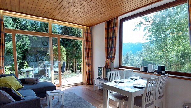 Ein modernes Wohnzimmer mit Sofa, Esstisch und Stühlen, großen Fenstern mit Vorhängen sowie Blick auf Grünflächen und Berge.