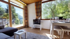 Ein modernes Wohnzimmer mit großem Fenster, einem Fernseher und einem Essbereich mit Stühlen und Tisch.