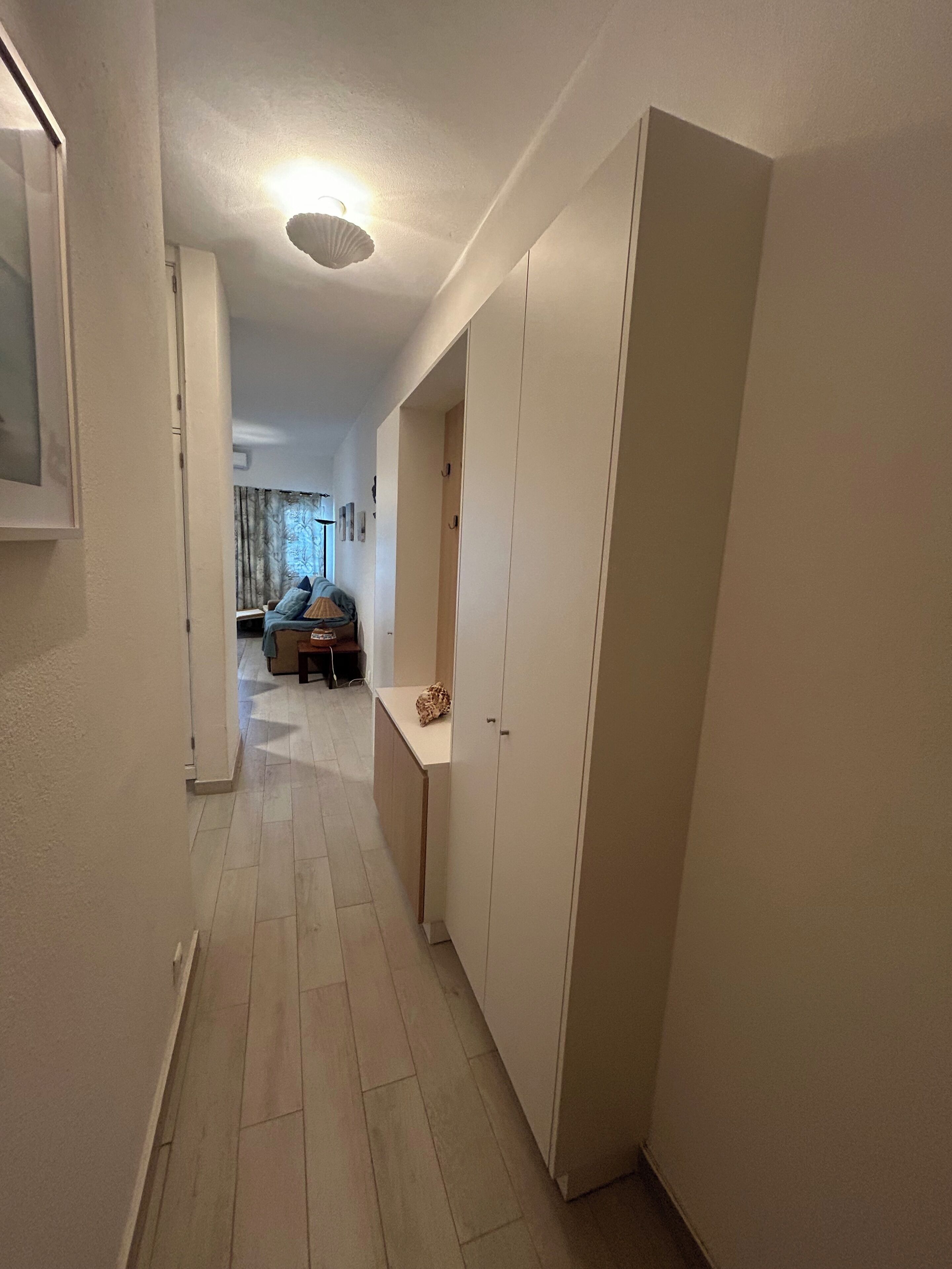 A narrow hallway with a light fixture on the ceiling, a door on the right, and a small table with a bag on it.
