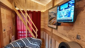 A cozy cabin room with a wooden interior, a red curtain, a patterned blanket on a sofa, and a television mounted on the wall.