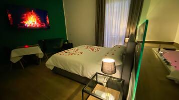 A hotel room with a bed, a TV mounted on the wall, a bedside table with a lamp, and a small table with a chair.