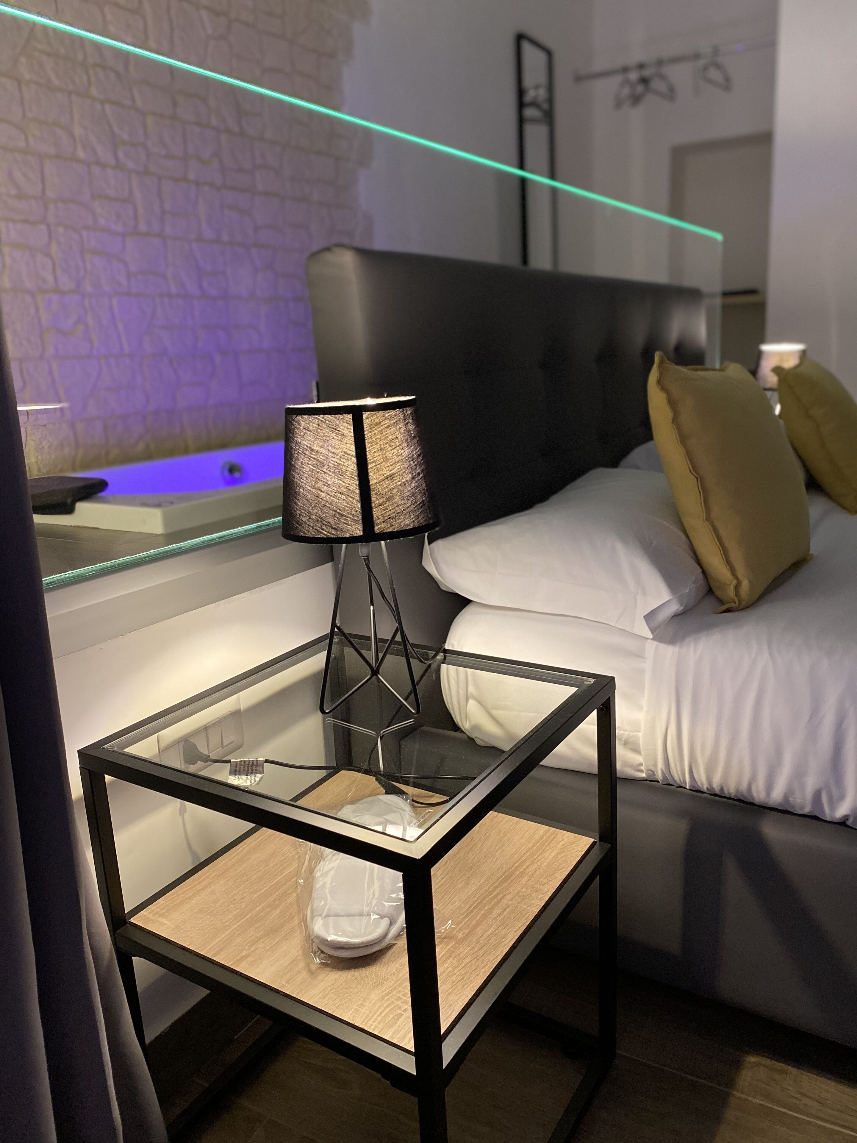 Quarto de hotel moderno com mesa lateral de vidro, abajur preto, cama com almofadas brancas e amarelas, e parede de tijolos.