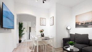 A modern living room with a black sofa, a coffee table with a plant, and a dining area with white chairs and a table. - Apartment close to Theresienwiese (München)