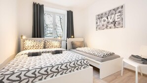 A modern bedroom with a large bed, a smaller bed, and a bench. There is a window with curtains, a wall-mounted artwork with the word "LOVE", and a chair. - Apartment close to Theresienwiese (München)