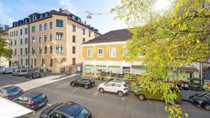 A street view with parked cars, a building with a yellow facade, and a tree with green leaves. - Apartment close to Theresienwiese (München)
