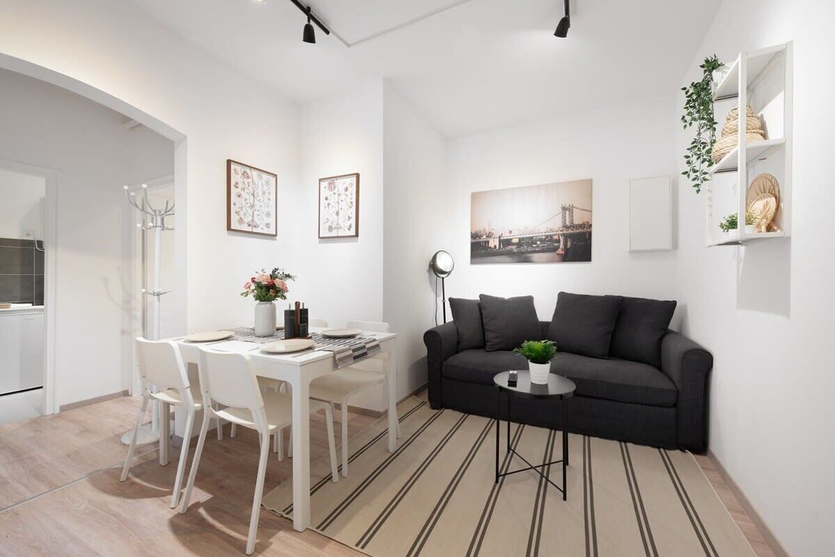 Ein modernes Wohnzimmer mit einer schwarzen Couch, einem weißen Esstisch und Stühlen. An den Wänden hängen gerahmte Bilder, und ein kleiner Beistelltisch trägt eine Pflanze.