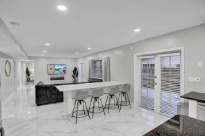 A modern kitchen with a white island, bar stools, and a dining area. - Incredible 7BR | Game Room Heated Pool and Jacuzzi (Deerfield Beach)