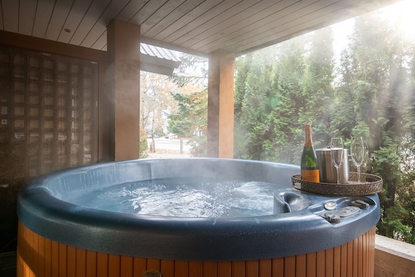 A hot tub with a bottle of champagne and glasses on a tray, surrounded by wooden panels and a view of trees.