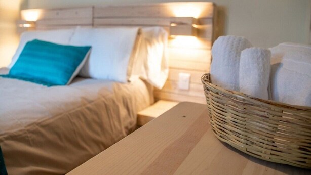 A neatly made bed with a blue pillow and a wooden nightstand beside it. A wicker basket filled with white towels is also visible. - Casa Cheleb (San Cristóbal de las Casas)