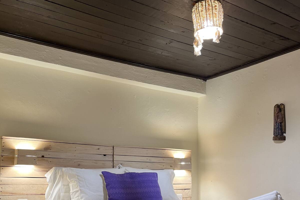 Ett sovrum med en sänggavel i trä, en säng med vita och lila sängkläder, ett nattduksbord med en lampa och en dekorativ detalj som hänger på väggen.