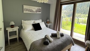 A bedroom with a large bed, a bedside table with a lamp, a window with a view of greenery, and a ceiling light fixture.