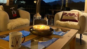 A cozy room with a wooden ceiling, a large window with a mountain view, two white armchairs, a table with a candle, and a lamp.