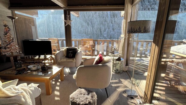 A cozy room with a wooden ceiling, a large window overlooking a snowy forest, a sofa, a chair, and a fluffy white animal.